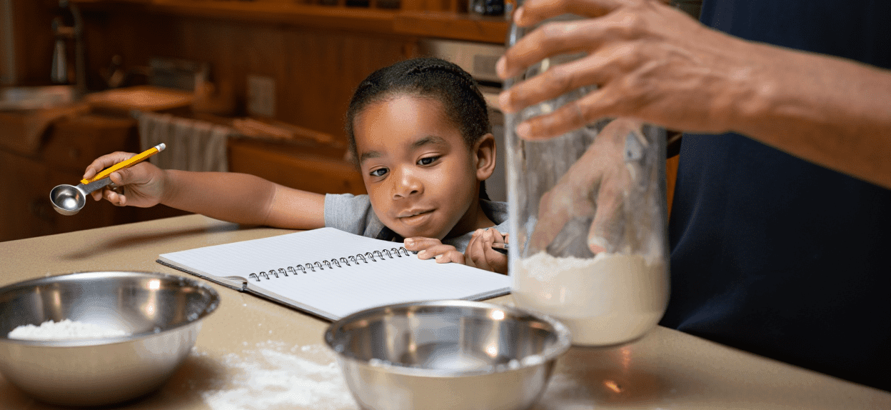 Child holding a measuring spoon, looking at a notebook, with an adult's hand in a flour jar.