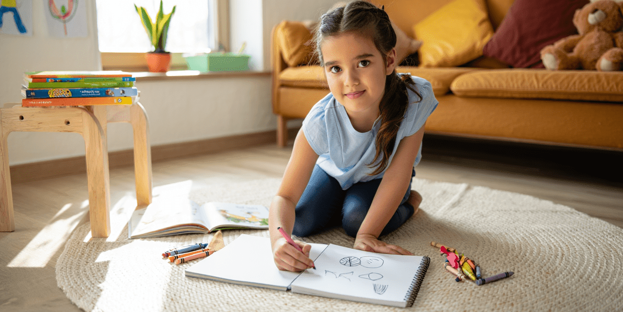 Child drawing in a sketchbook on the floor with crayons nearby.
